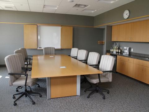 Room with large conference table surrounded by comfortable arm chairs, a cabinet with white board, and a counter with a sink and cabinets