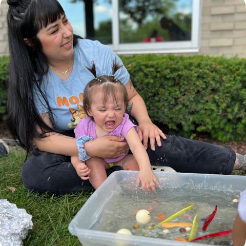 Baby sitting in mom's lap and splashing in a tub of water