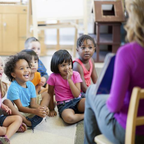 Children sitting smiling while listening to an adult read a book in the foreground