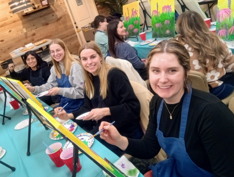 Young ladies sitting at easels with paint brushes and looking at the camera