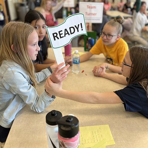 Four girls sitting around a table holding hands with one girl holding up a sign that says "Ready!"